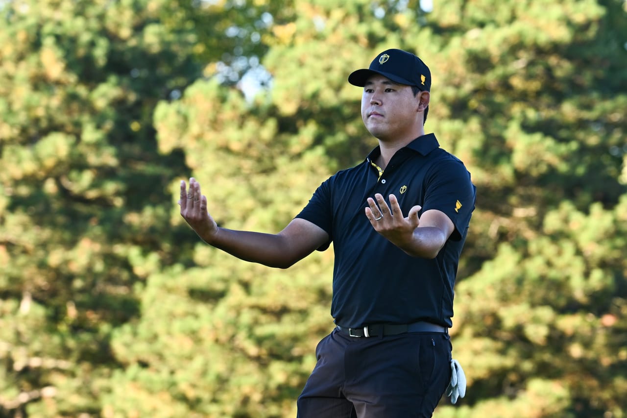 Si Woo Kim, in International team black and gold, throws both hands out toward the gallery in front of a wall of pines