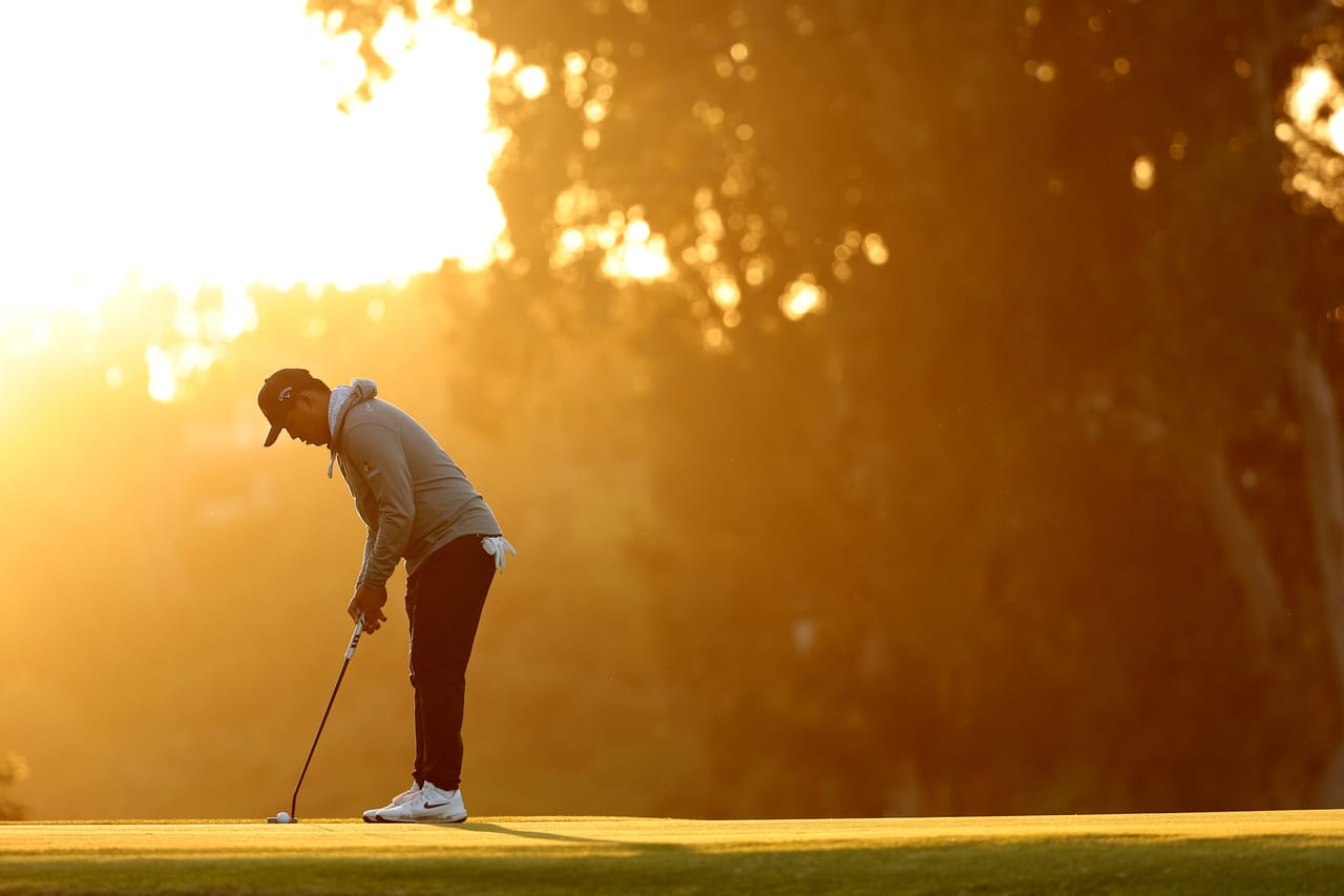 Si Woo Kim, alone on a green, stands over a putt as a low sun burns gold through the trees behind him